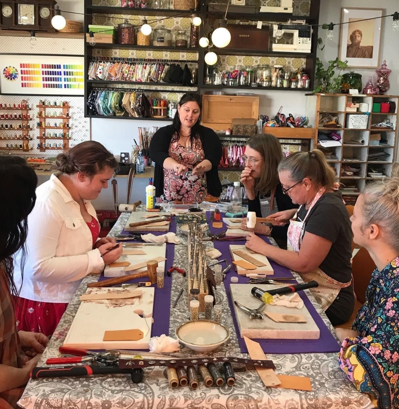 Picture of a leatherwork class in a craft room with five participants and one teacher standing around a table